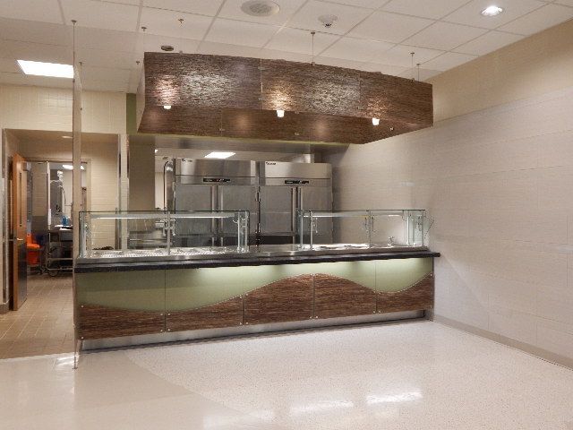 A cafeteria serving station with a wooden-textured overhead canopy and base, glass sneeze guards, and stainless appliances.