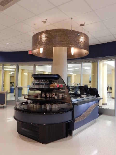 A tiered, curved refrigerated display case for food and drinks stands in an indoor space with a decorative drum light.