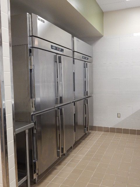 Two large stainless-steel commercial refrigerators stand side-by-side against a white-tiled wall in a kitchen area.