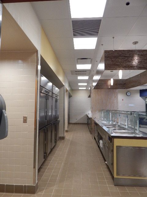 A narrow cafeteria kitchen aisle with tall stainless steel refrigerators on the left and a service counter on the right.