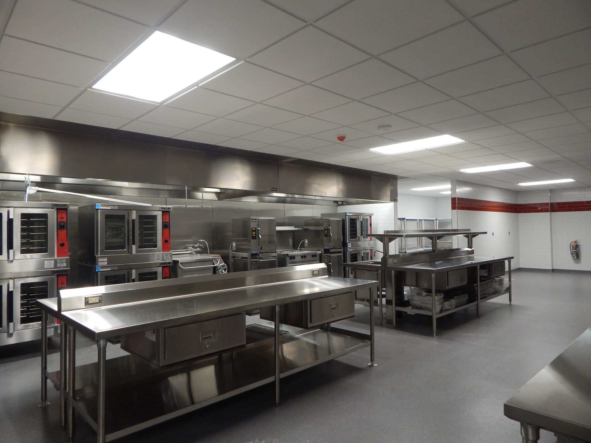 A commercial kitchen featuring stainless steel work tables, rows of ovens, and a drop ceiling with fluorescent lighting.