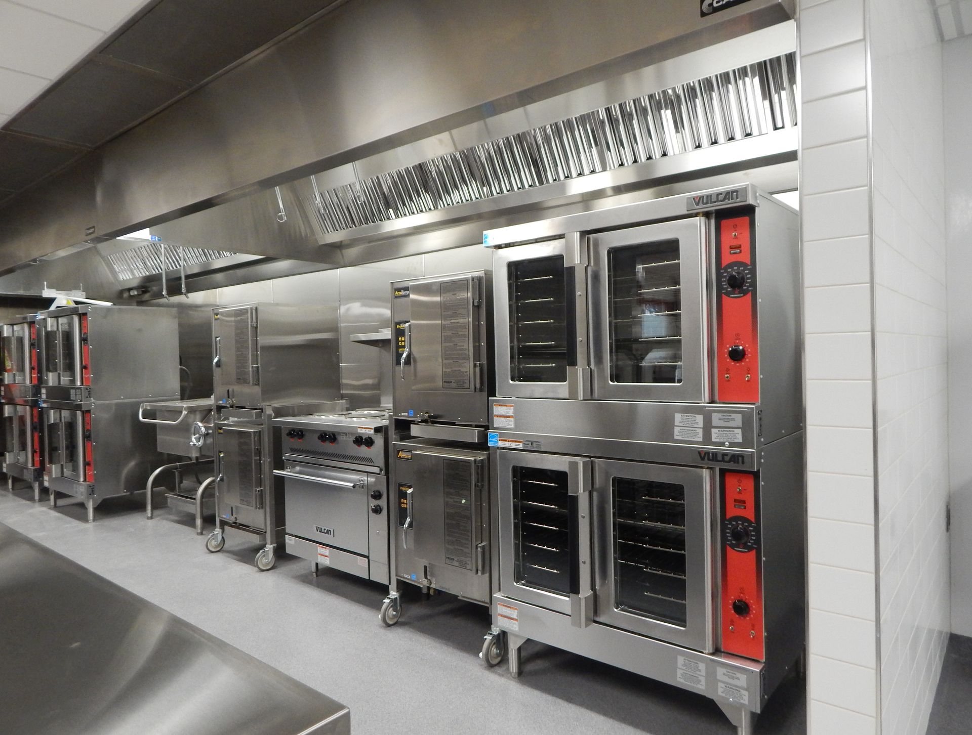 A row of stainless steel commercial kitchen appliances, including stacked convection ovens, situated under a ventilation hood.