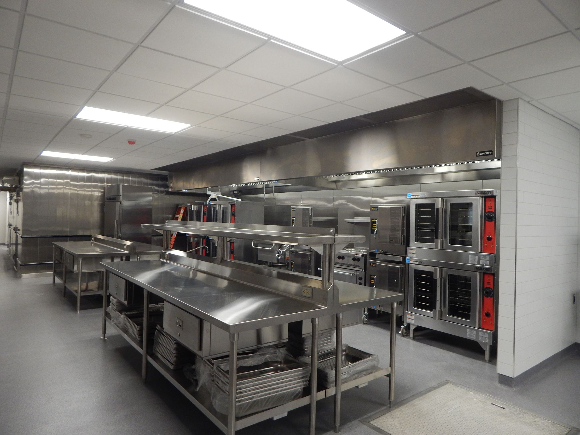 A modern commercial kitchen with stainless steel work tables, large ovens, and ventilation hoods under bright lights.