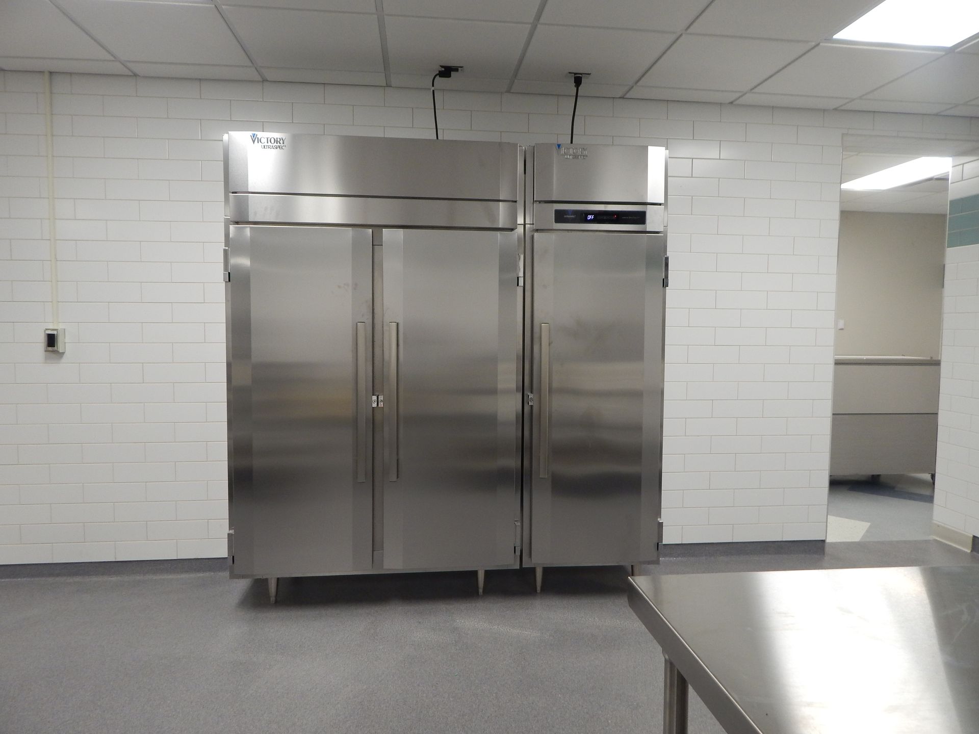 Two stainless steel commercial refrigerators stand against a white brick wall in a kitchen.