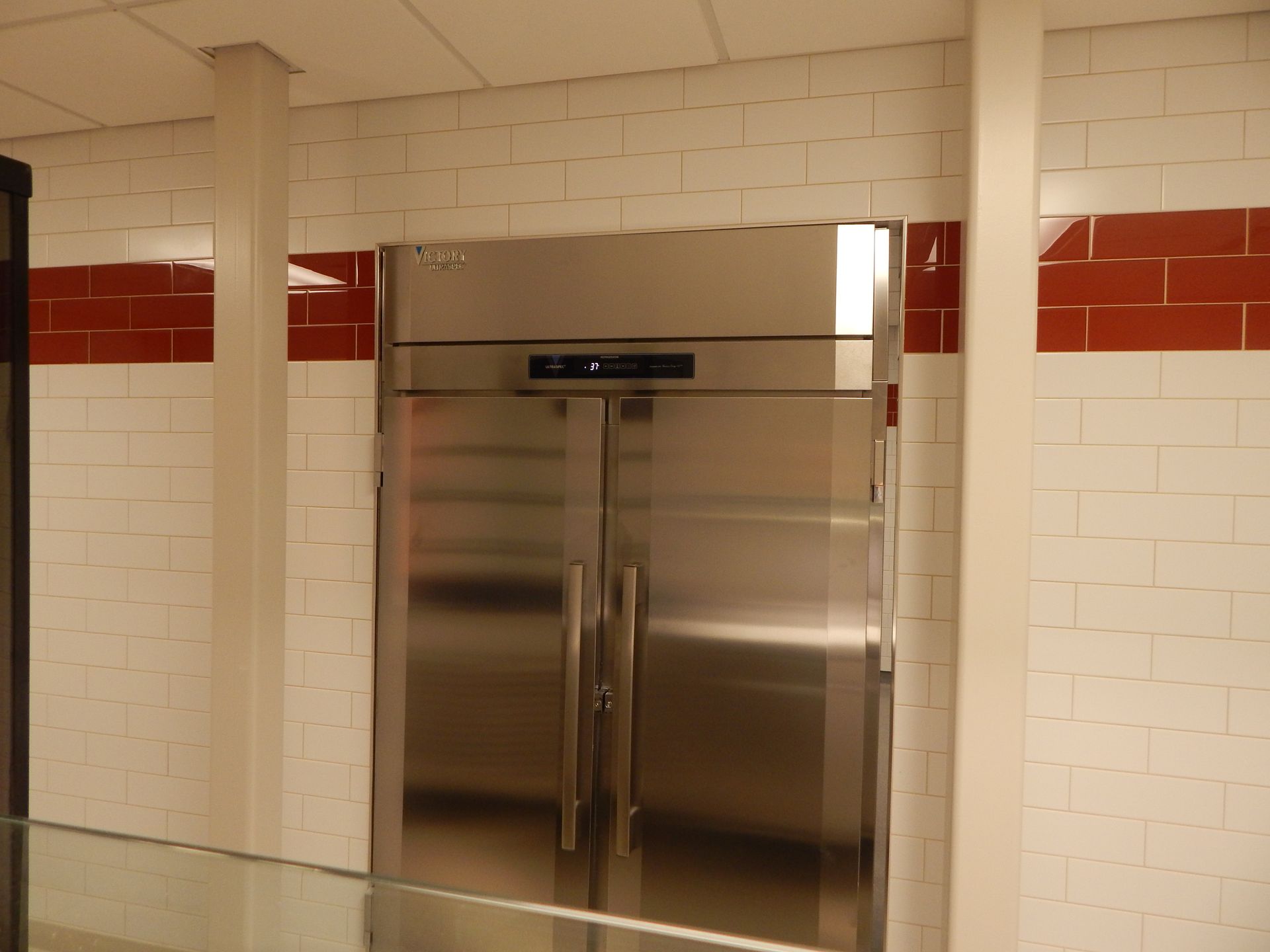 A large, stainless steel, double-door commercial refrigerator set into a white-tiled wall with a horizontal red stripe.