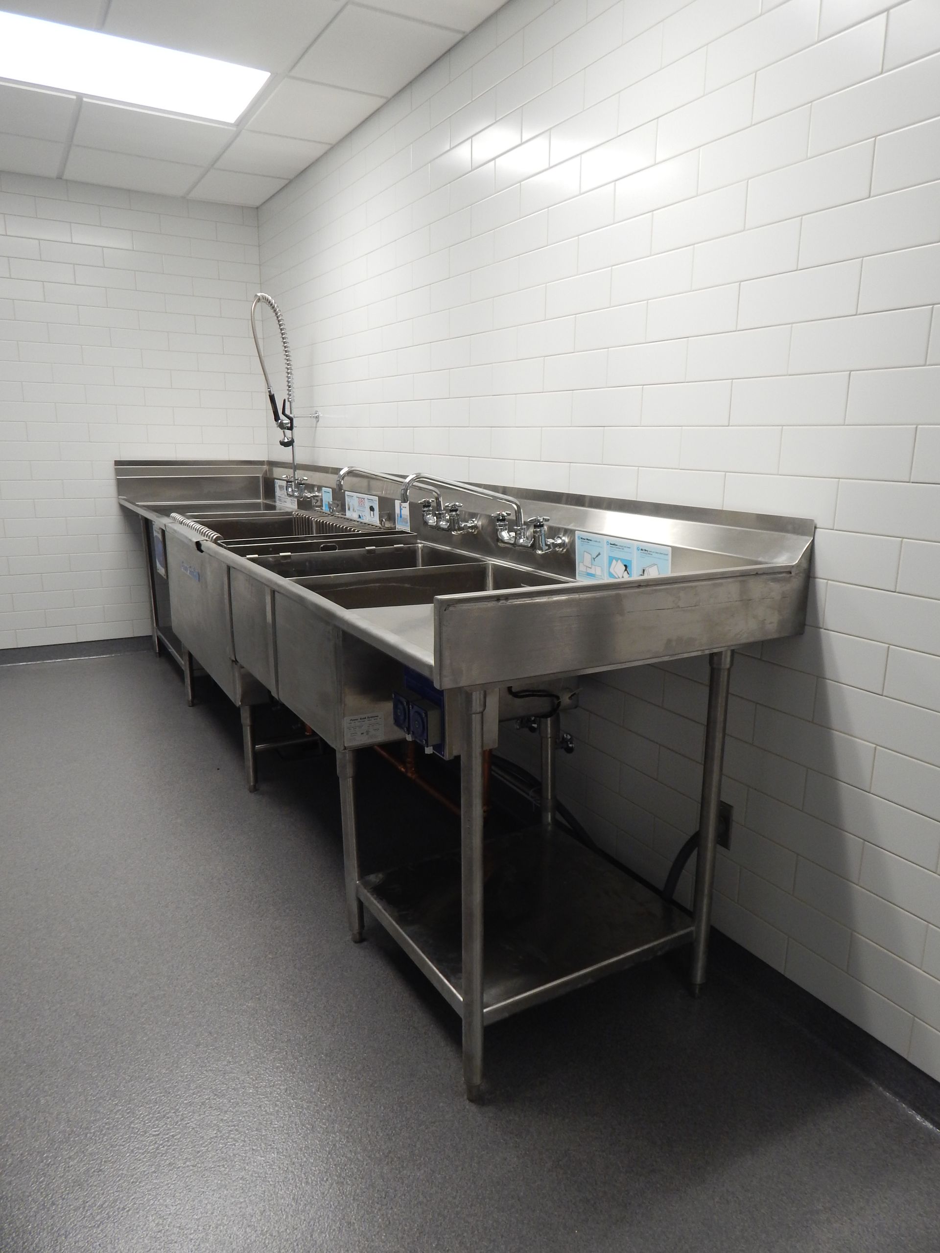 A long, stainless steel multi-compartment sink against a white-tiled wall in a commercial kitchen setting.