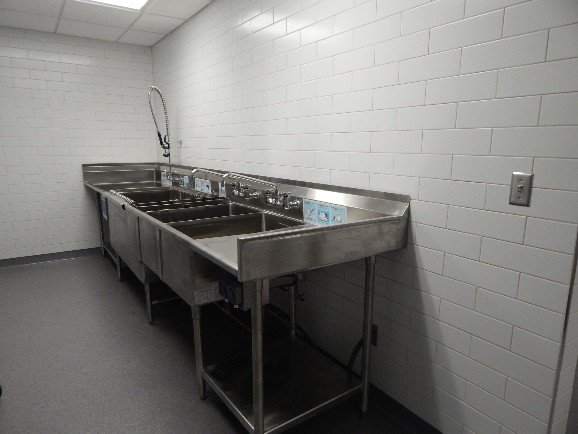 A stainless steel three-compartment industrial sink with a sprayer in a commercial kitchen with white tiled walls.