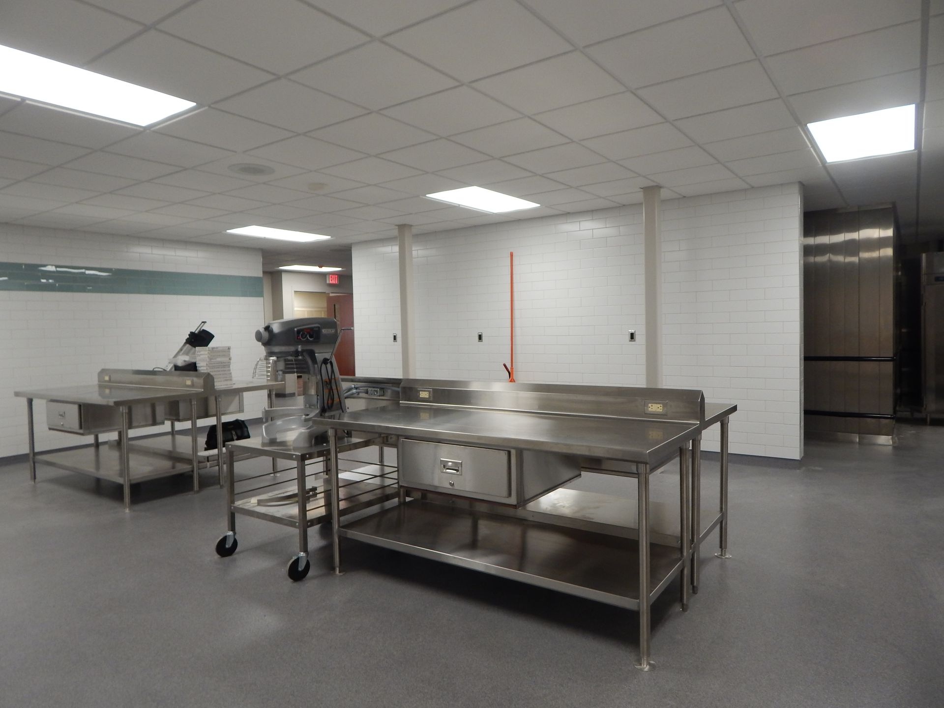 A commercial kitchen with stainless steel tables, a floor mixer, and a white tile wall under bright fluorescent lighting.