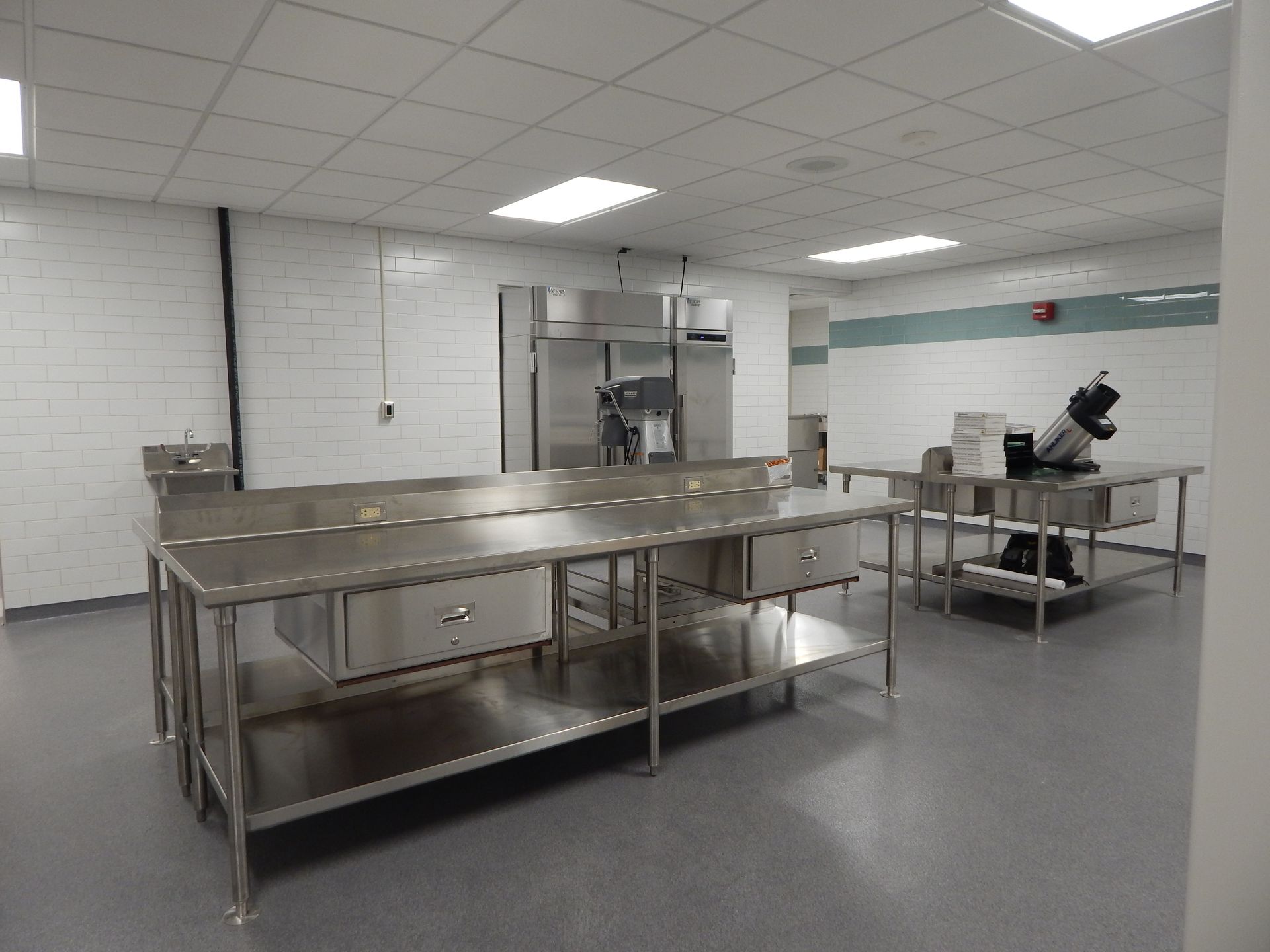 A sterile, white-walled commercial kitchen featuring stainless steel prep tables and a commercial mixer.