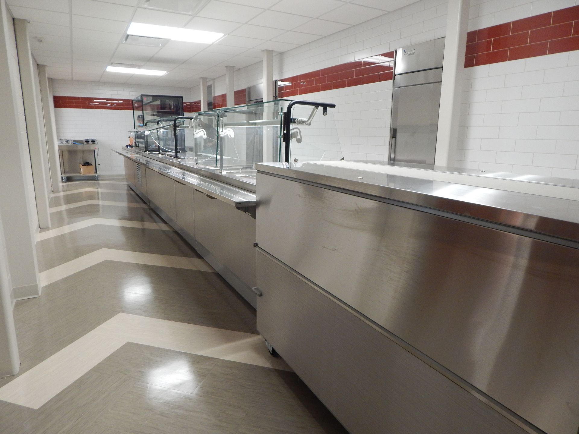 A long, stainless steel cafeteria serving line with glass sneeze guards in a room with a chevron-patterned floor.