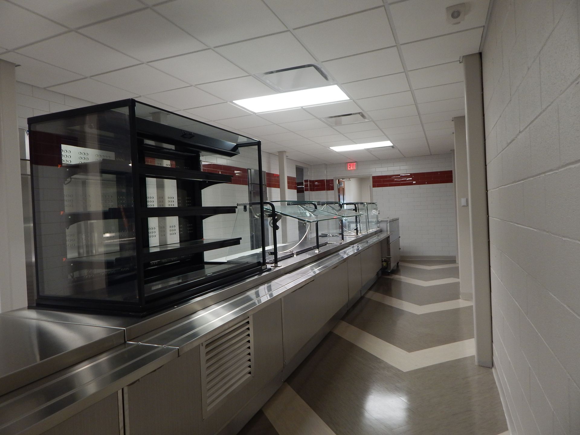 A metal food service counter with glass display shelving, set in a hallway with white block walls and chevron floor tiles.