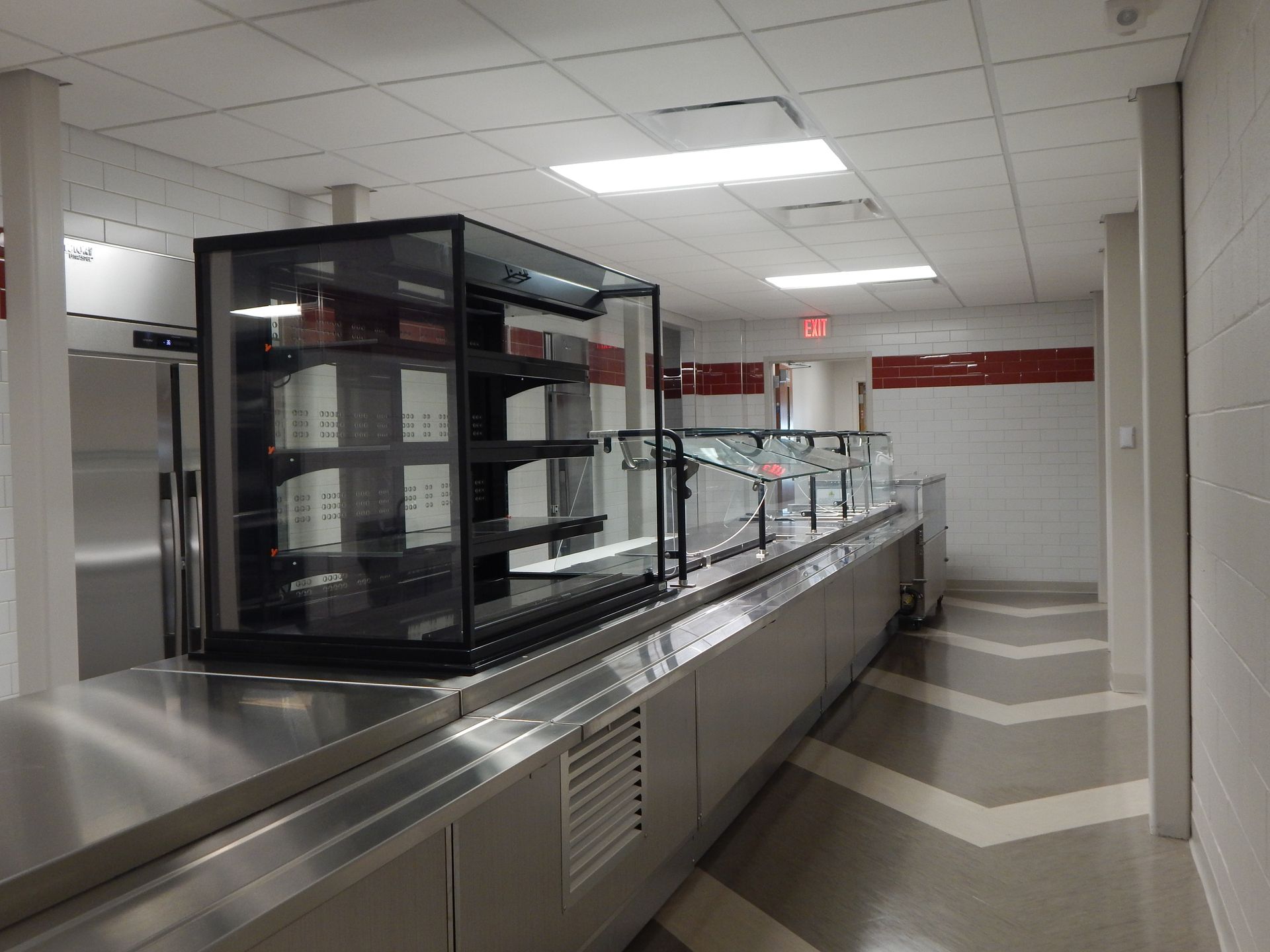 A stainless steel food service counter with a display case in a modern, brightly lit institutional cafeteria hallway.