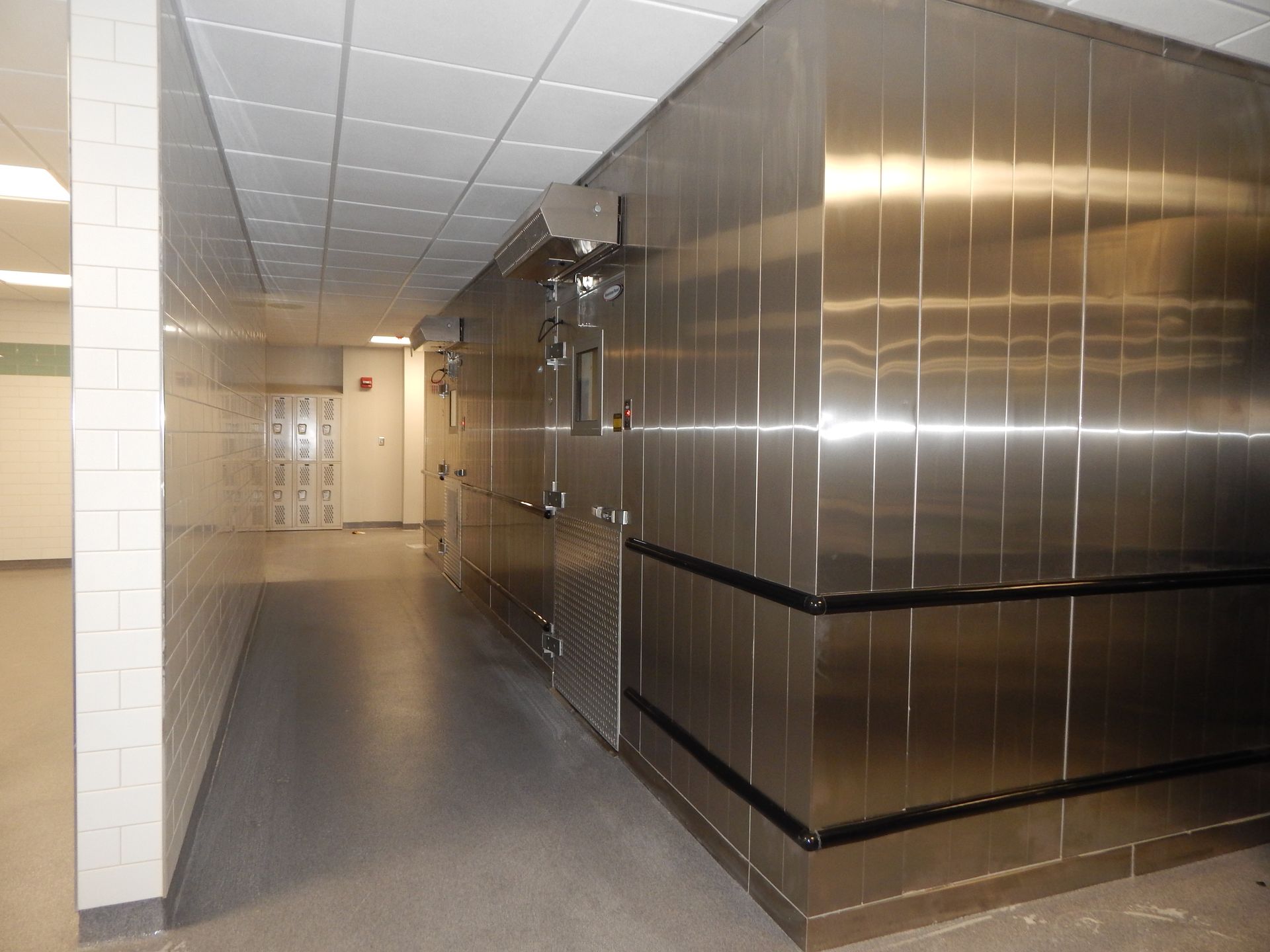 A narrow corridor with white tiled walls on the left and a large, stainless steel industrial cooler on the right.