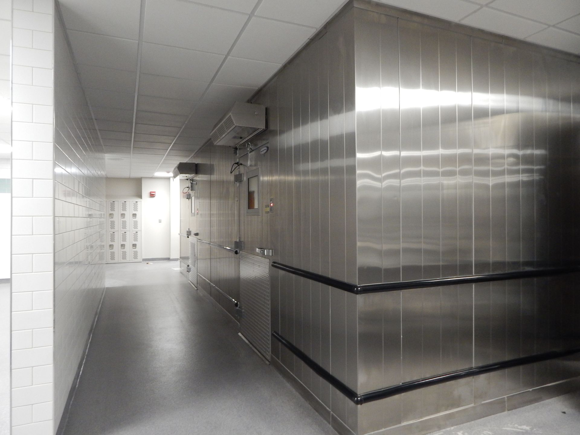 A long hallway with a white-tiled wall on the left and a large, stainless steel walk-in cooler on the right.