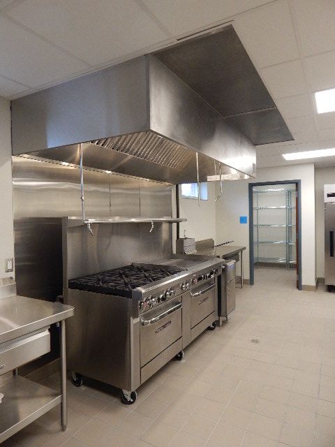 Commercial kitchen with a stainless steel range and large exhaust hood, tiled floor, and an open doorway in the background.
