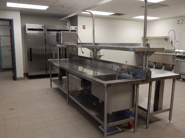 A commercial kitchen featuring stainless steel prep tables, a sink, an overhead shelf, and large metal refrigerators.