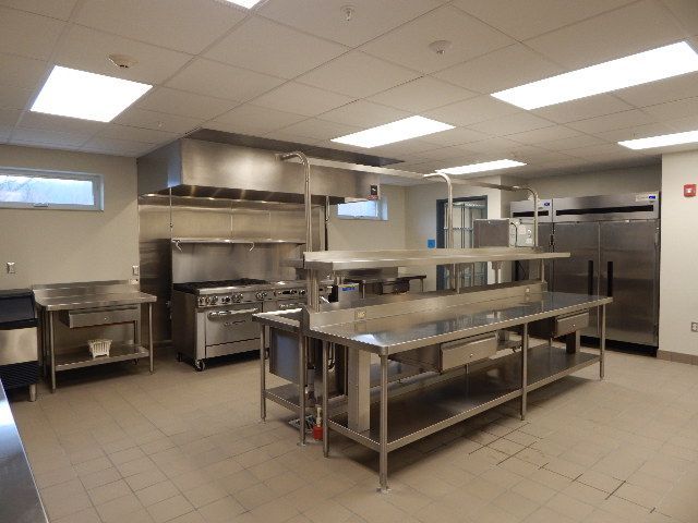 A bright, clean professional kitchen featuring stainless steel food prep tables, a large stove, and a commercial fridge.