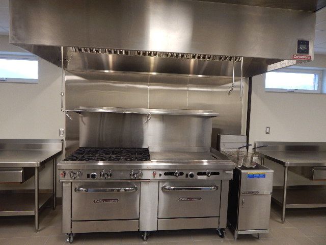 Commercial kitchen with a stainless steel range, oven, hood vent, and preparation counters in a room with white walls.
