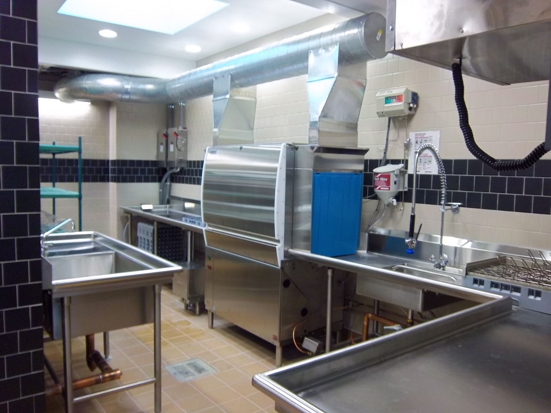 Commercial dishwashing area featuring a stainless steel dishwasher, metal sinks, and ventilation ducts in a tiled room.