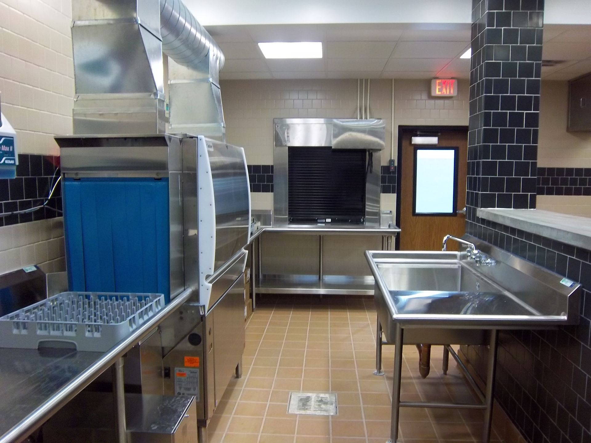 Commercial kitchen dishwashing area with a metal dishwasher, stainless steel sink, and black tiled walls.