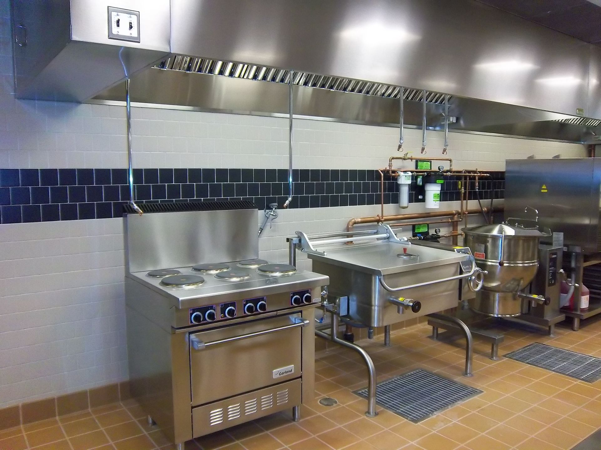 A commercial kitchen featuring stainless steel appliances, including a stove, tilt skillet, and large kettle under a vent.