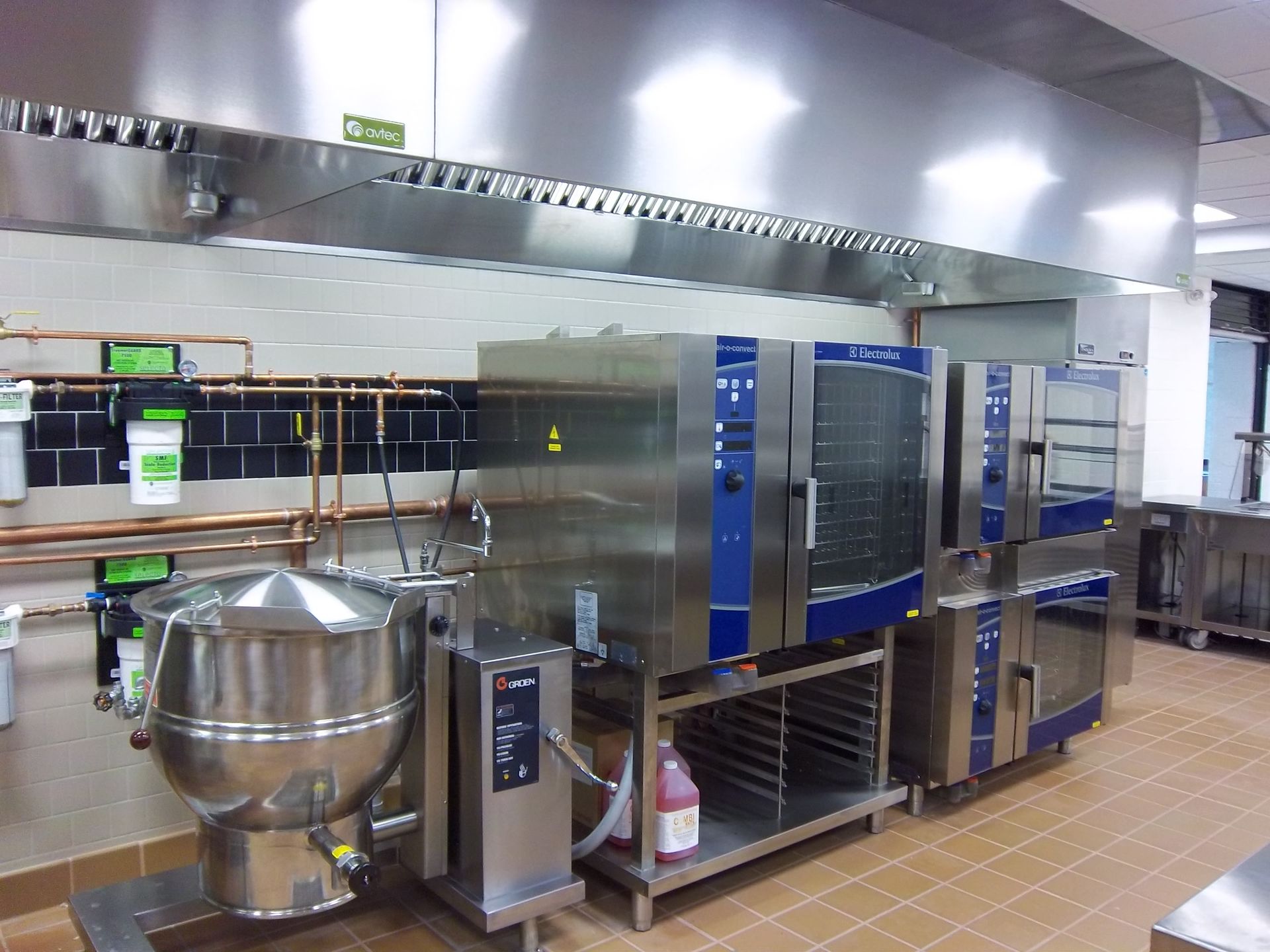 Commercial kitchen featuring a large stainless steel tilting kettle and stacked industrial ovens under a metal hood.
