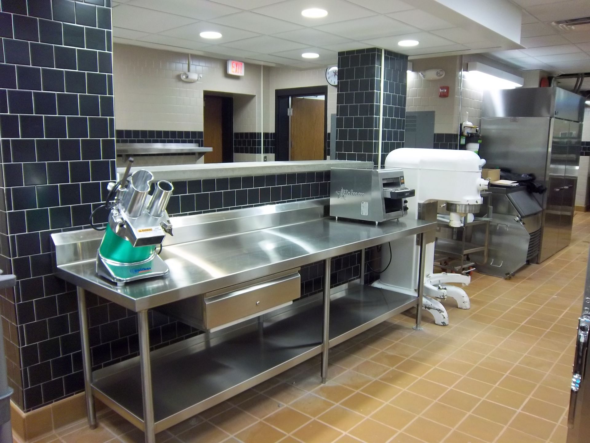 A professional kitchen features a long stainless steel prep table, a food slicer, and heavy-duty appliances on tiled floors.