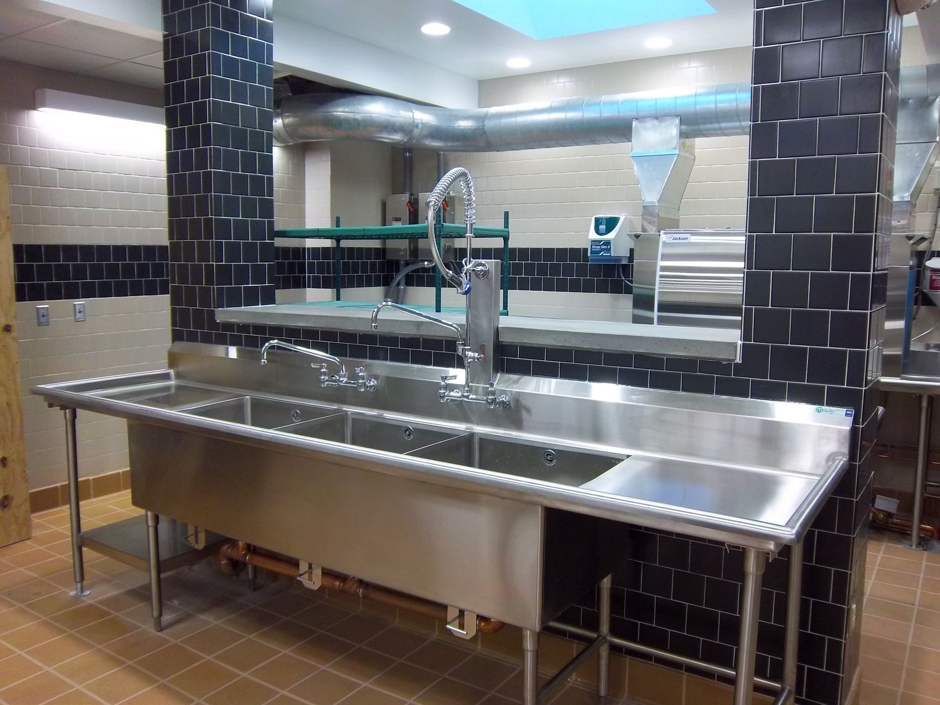 A large, stainless-steel, three-compartment commercial sink set between two black-tiled pillars in a kitchen.