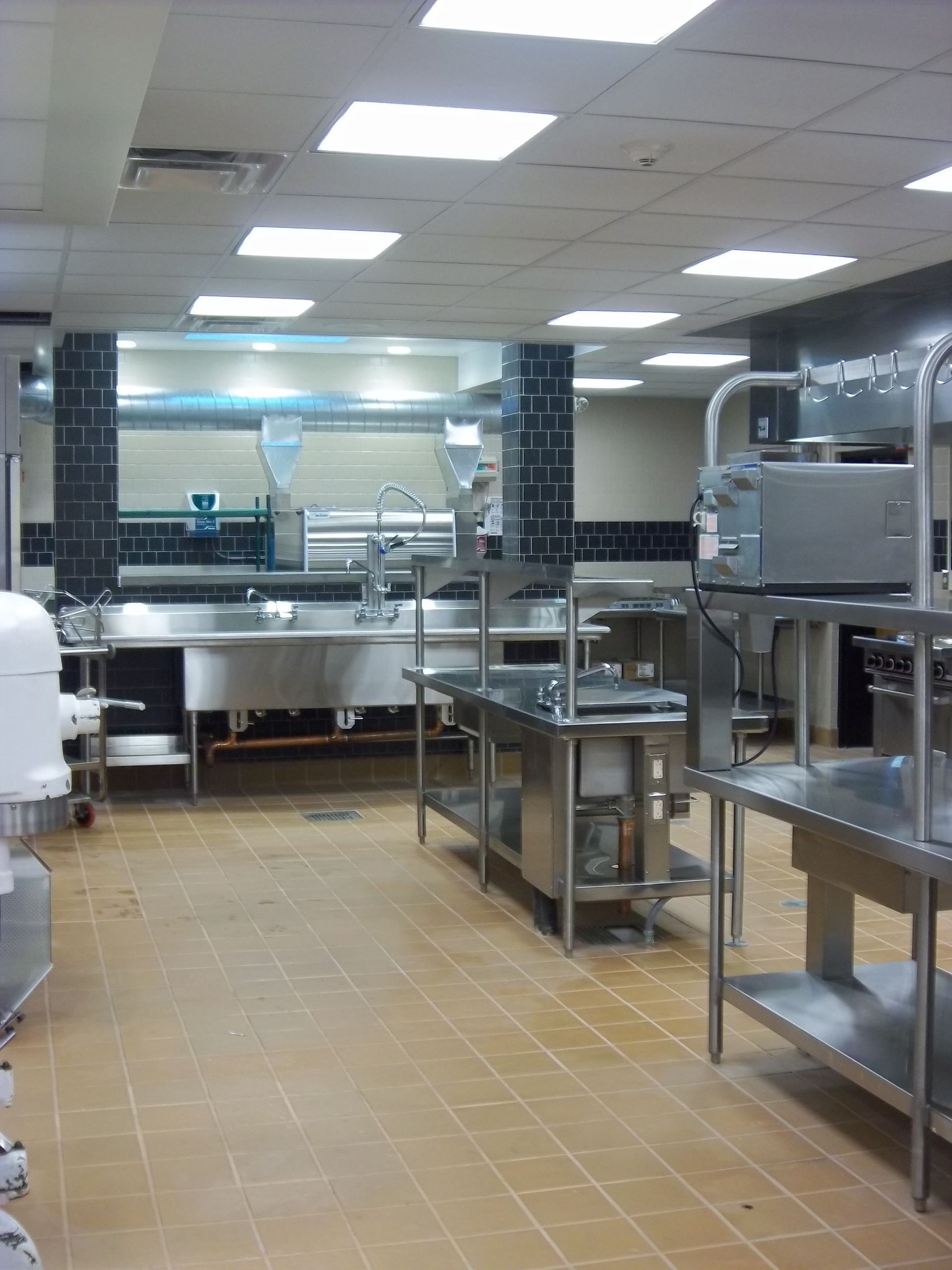 An empty, professional industrial kitchen with stainless steel tables, counters, a sink, and square ceiling lights.