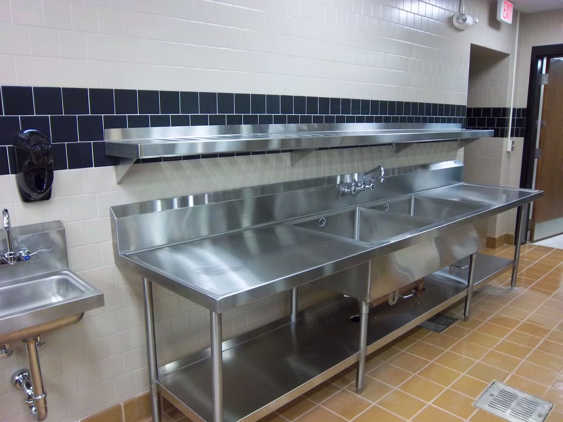 A commercial kitchen featuring stainless steel sinks, prep tables, and shelves against tiled walls with an orange floor.