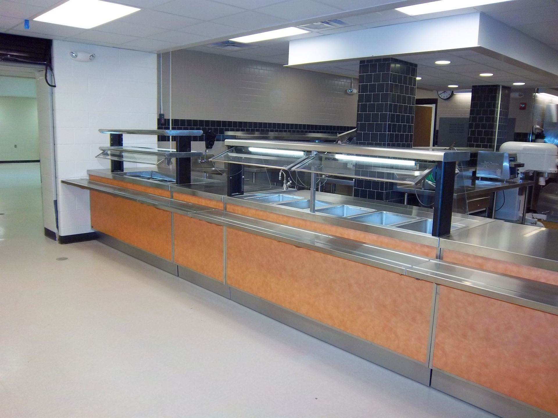 An empty cafeteria serving line with a light wood-tone front, stainless steel counter, and overhead sneeze guards.