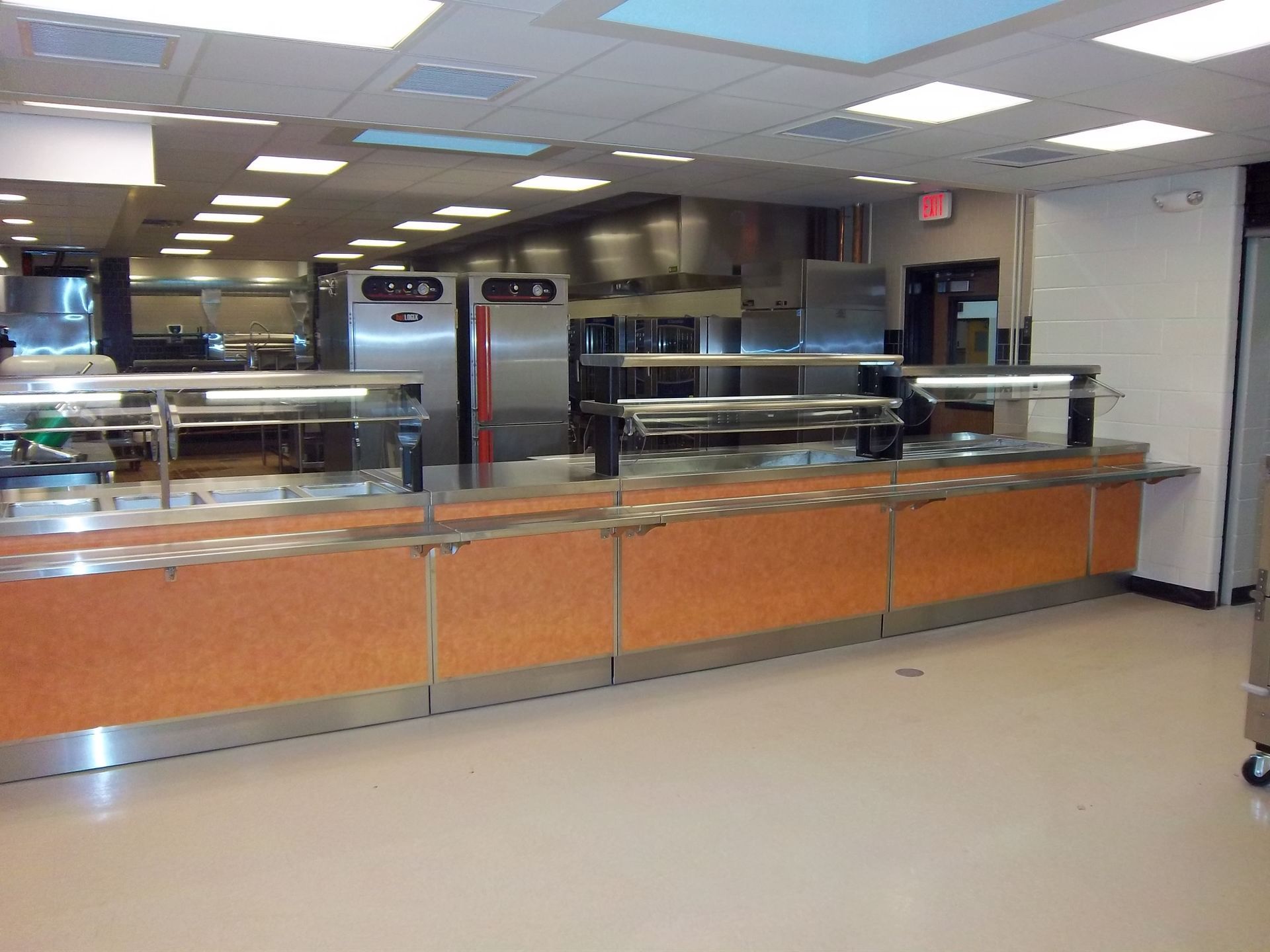 A commercial cafeteria serving line with stainless steel food warmers and orange base cabinets in a brightly lit kitchen.