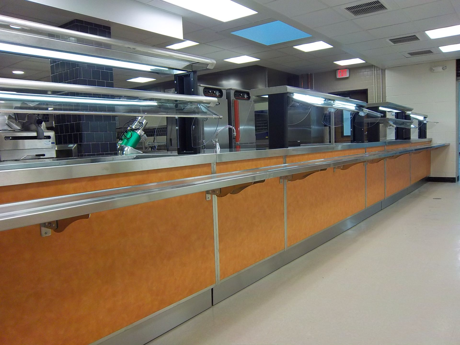 A long, empty cafeteria serving line with orange wooden panels, stainless steel countertops, and overhead lighting.