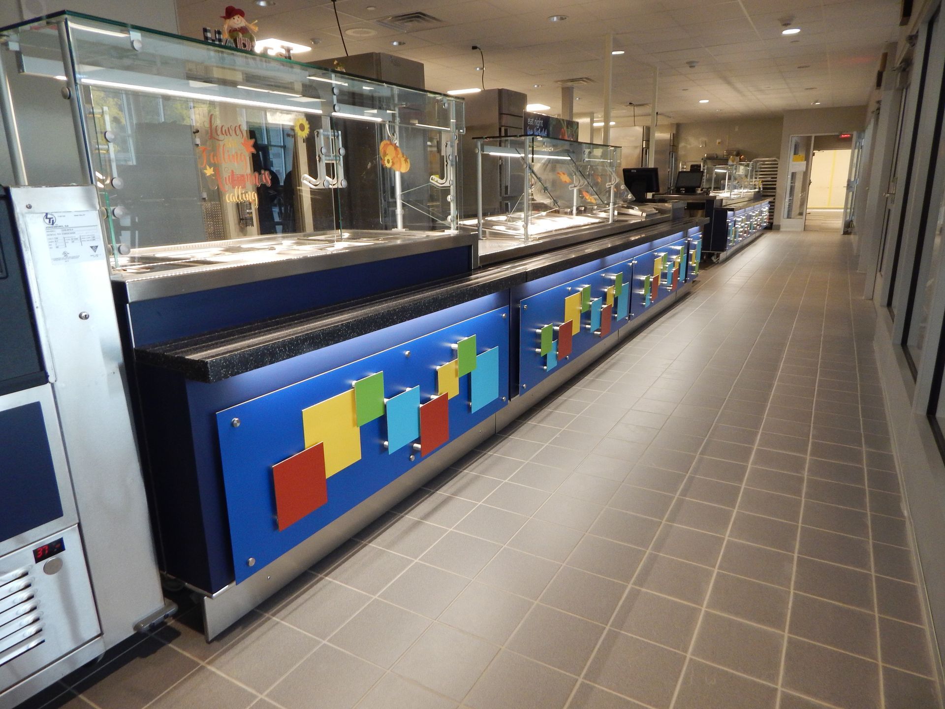 A long, cafeteria-style serving counter with blue panels decorated with colorful square accents on a tile floor.