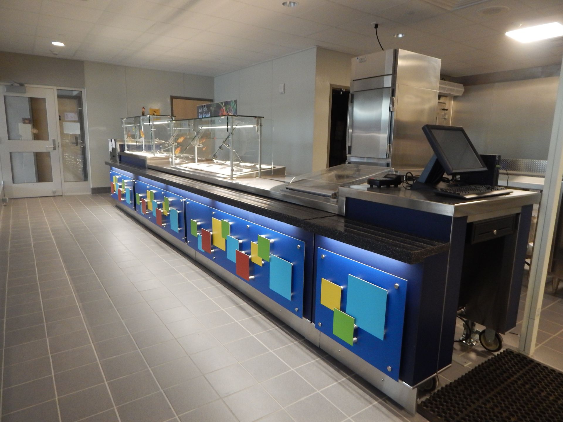 A food service counter with blue panels decorated with colorful square accents, located in a tiled, well-lit cafeteria.