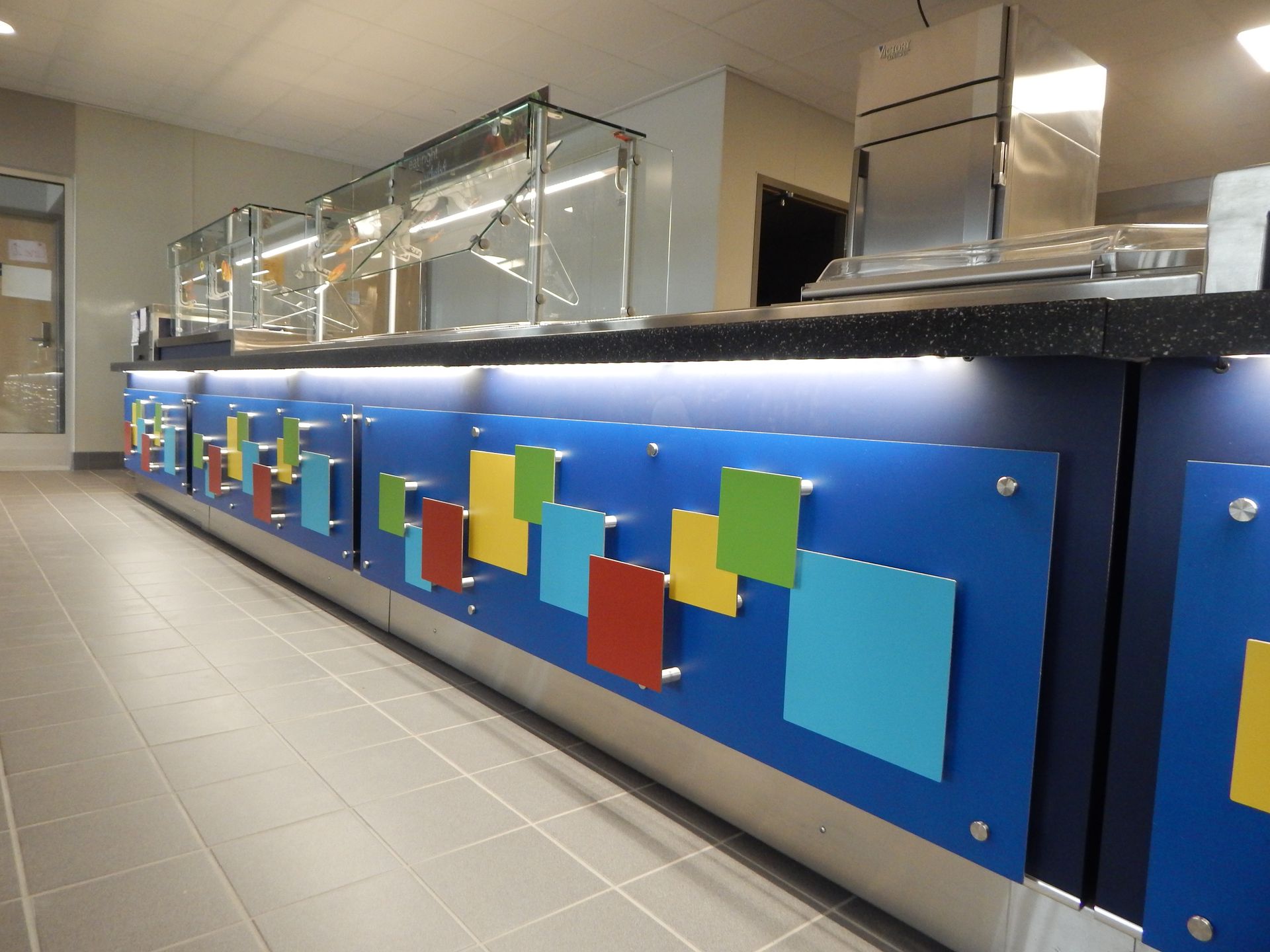 A serving counter with a blue front decorated with colorful square accents and a glass sneeze guard above.