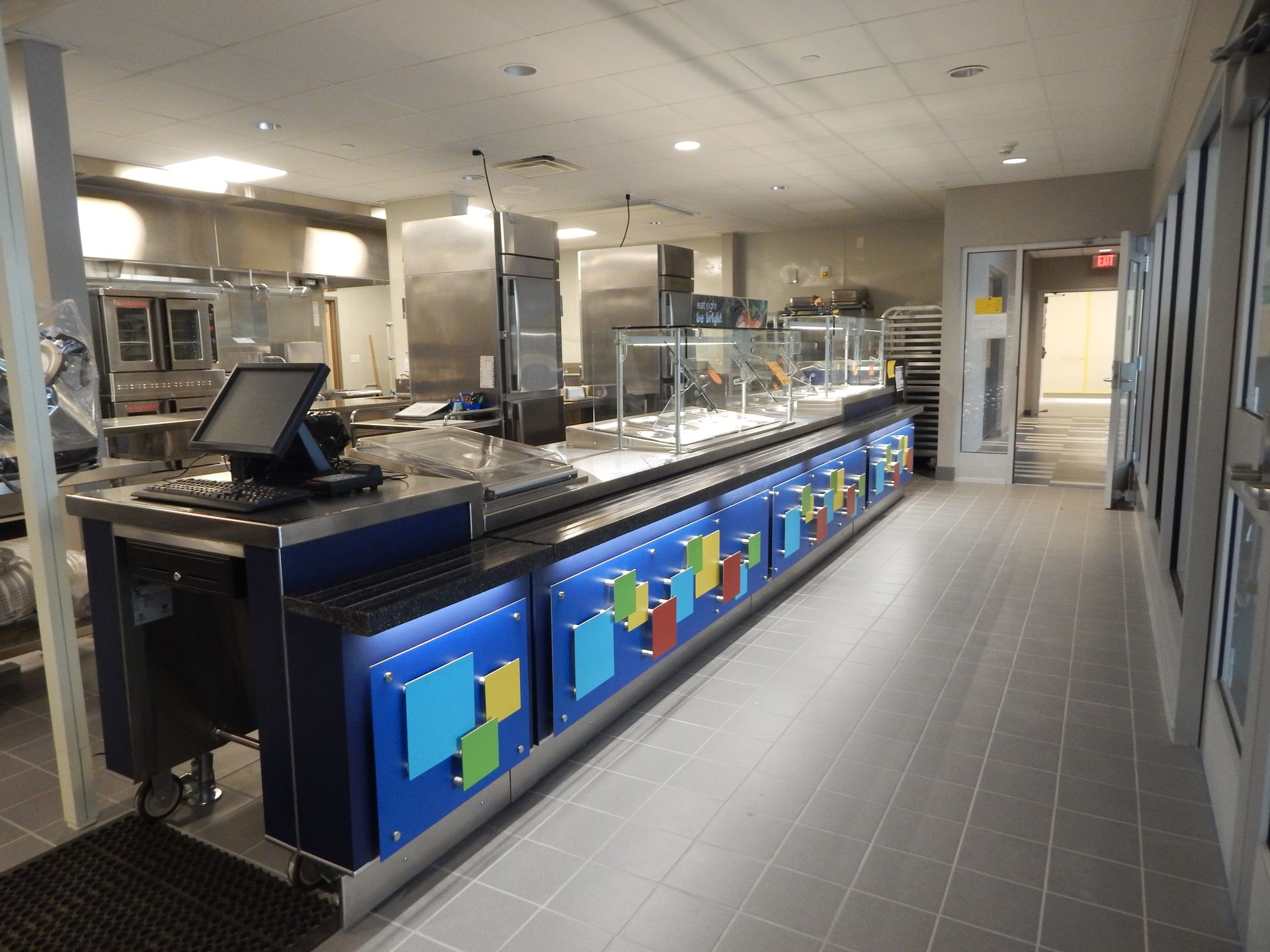 A commercial kitchen food service counter with blue panels featuring colorful geometric shapes, set in a tiled room.