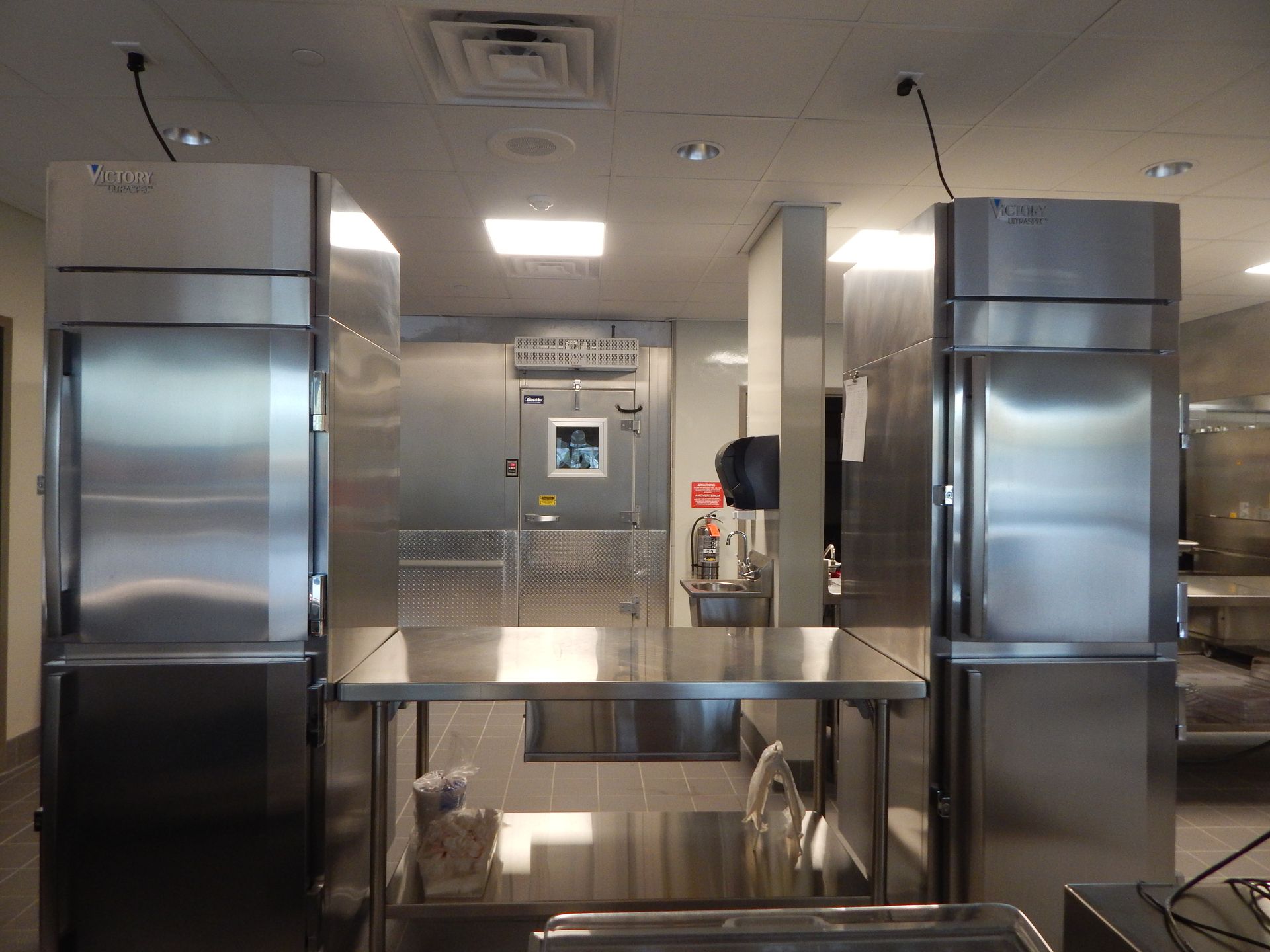 A stainless steel commercial kitchen featuring two upright refrigerators flanking a metal prep table.