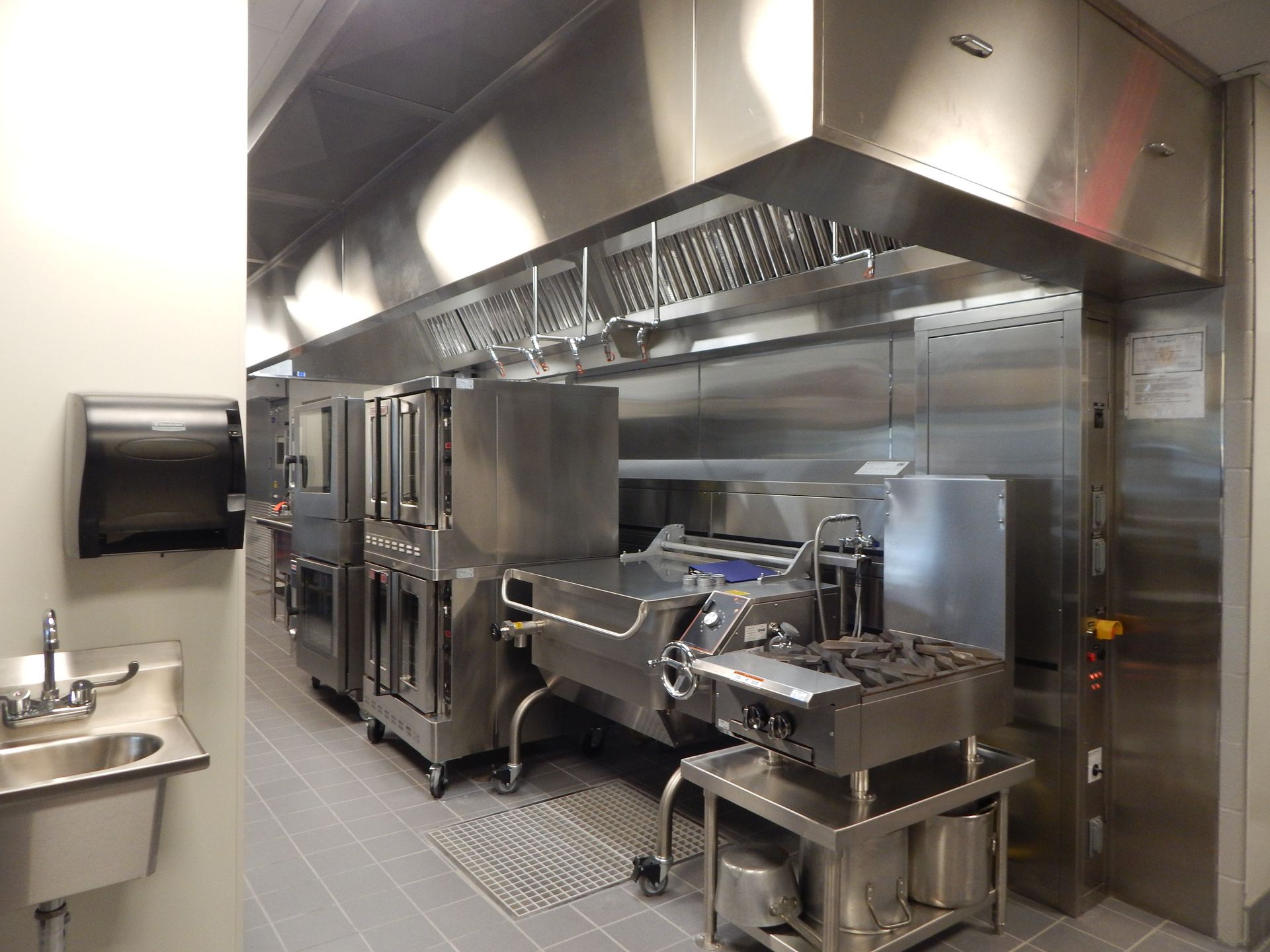 A stainless steel commercial kitchen featuring ovens, a tilt skillet, a stovetop, and a ventilation hood above.