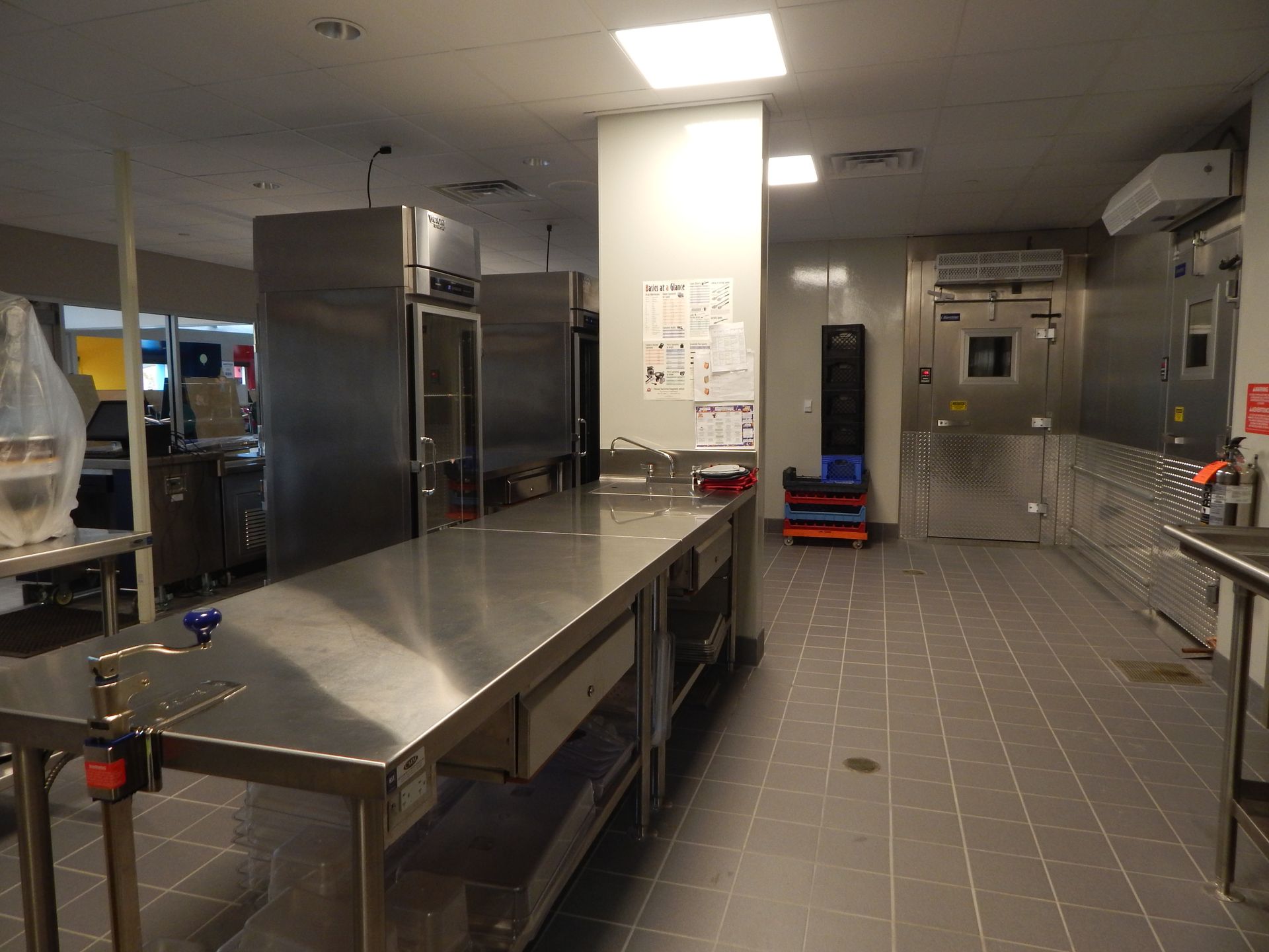 A wide-angle view of a commercial kitchen featuring stainless steel tables, refrigerators, and tiled flooring.