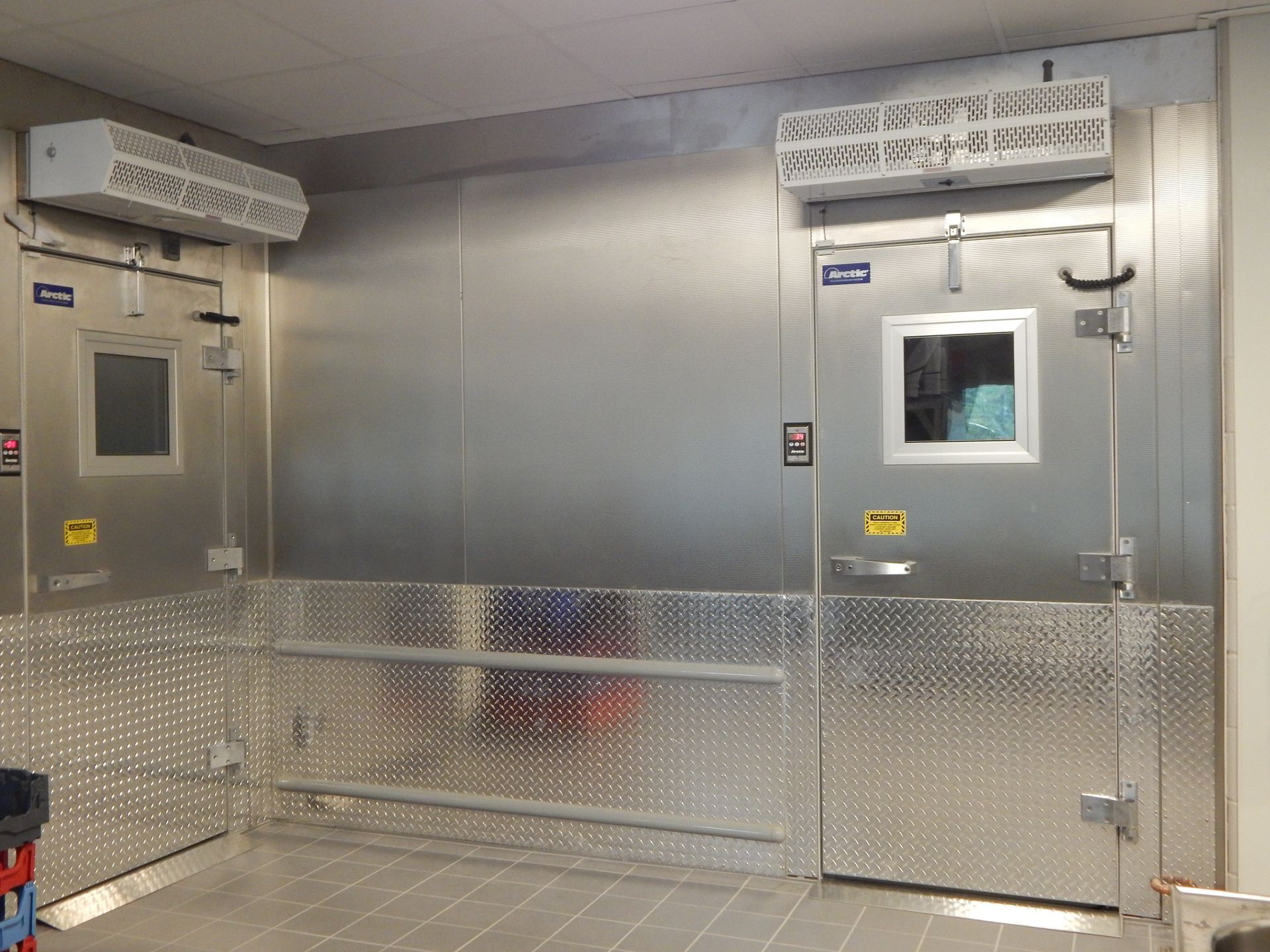 Two industrial walk-in coolers with metal doors, glass windows, and overhead refrigeration units in a tiled room.