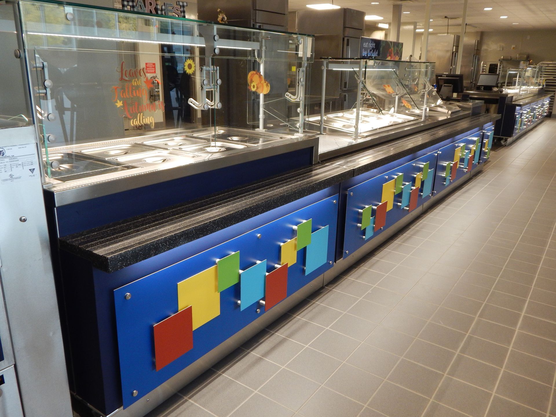 A long buffet food serving counter with blue panels decorated with colorful square accents in an industrial cafeteria.
