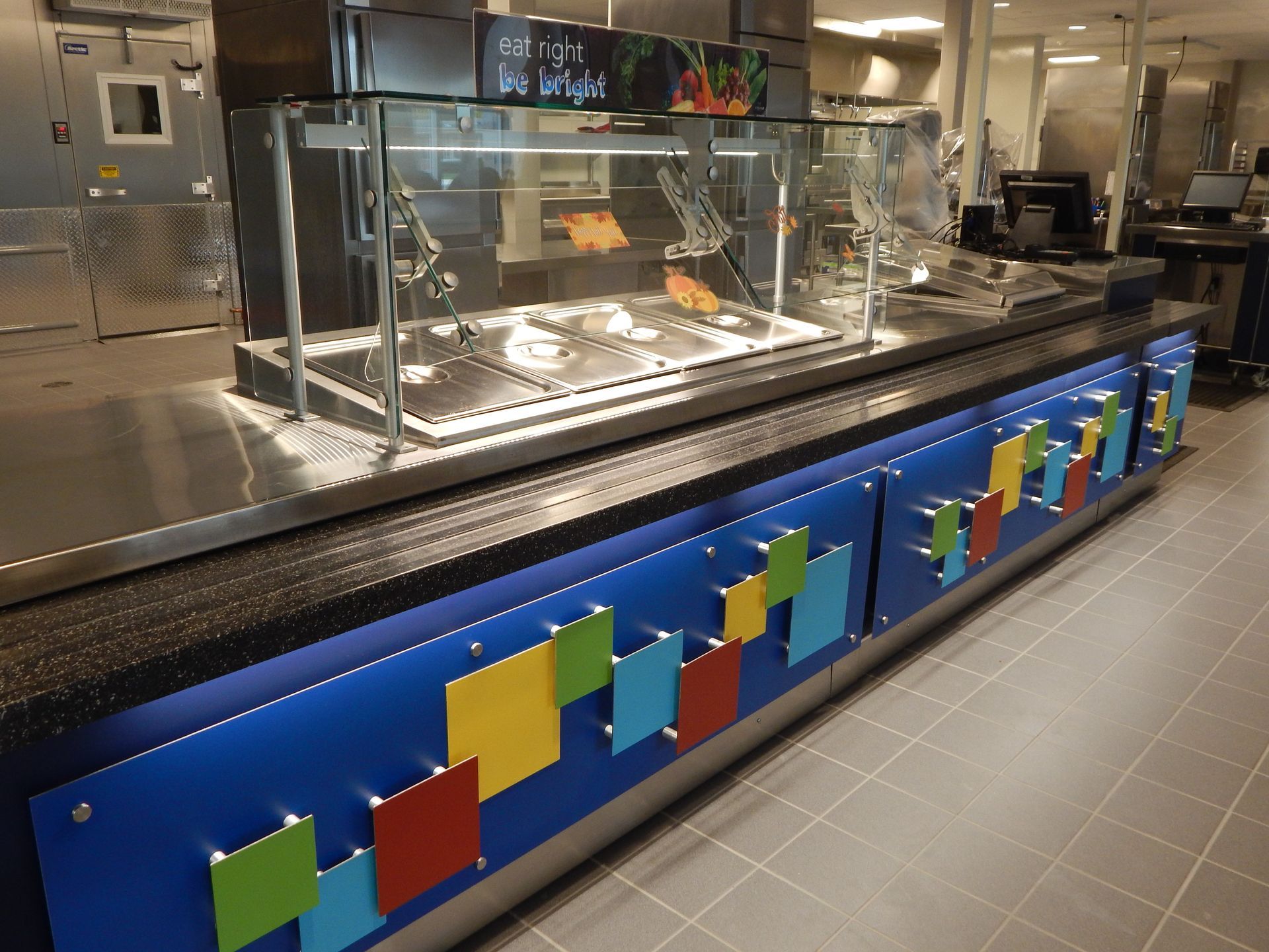 A cafeteria serving station with food warmers, a black countertop, and a blue base decorated with colorful geometric tiles.
