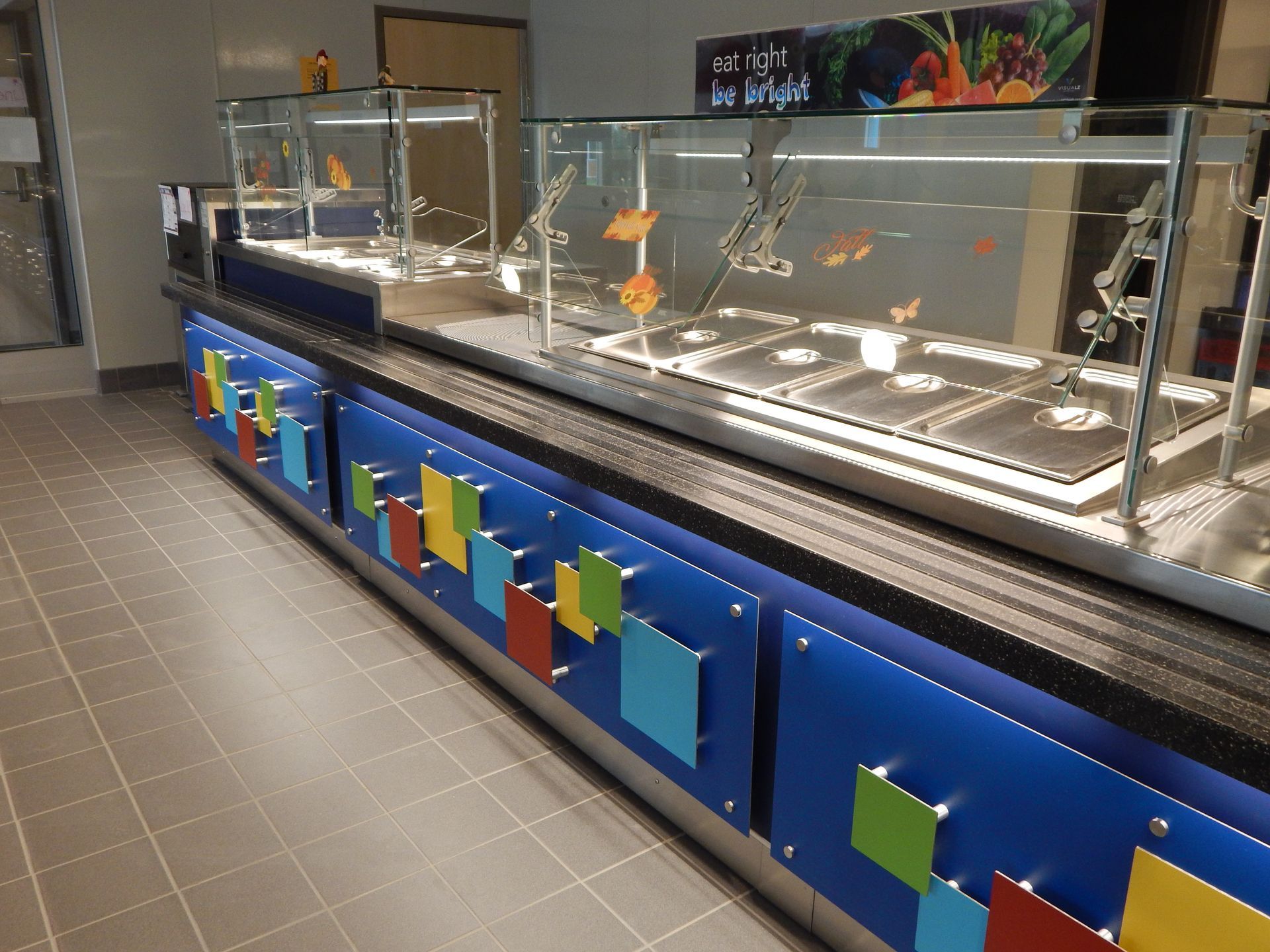 A cafeteria serving line with a glass sneeze guard and colorful square accents on a blue counter.