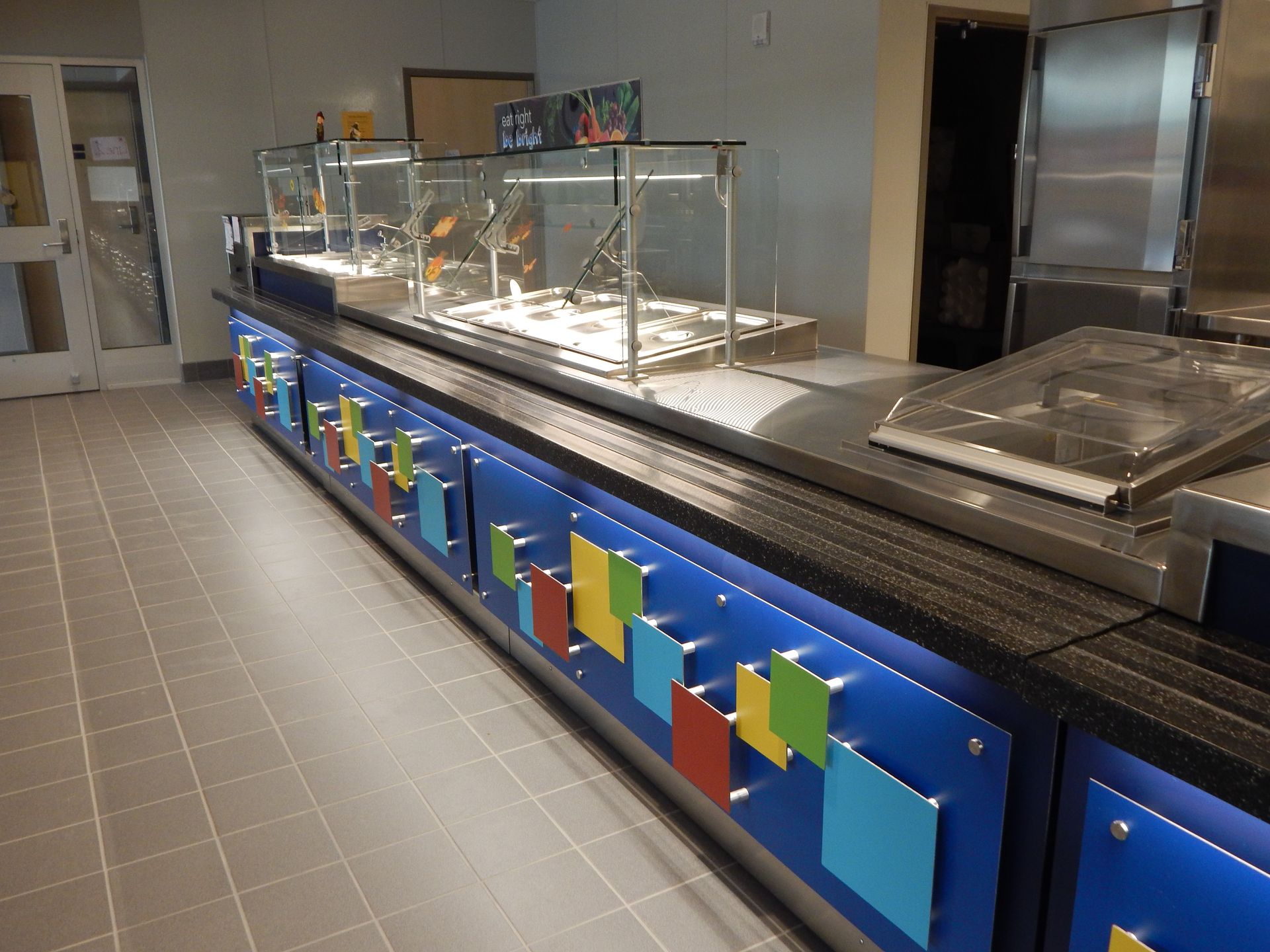 A buffet serving counter with a blue front decorated with colorful square accents, located in a tiled cafeteria area.