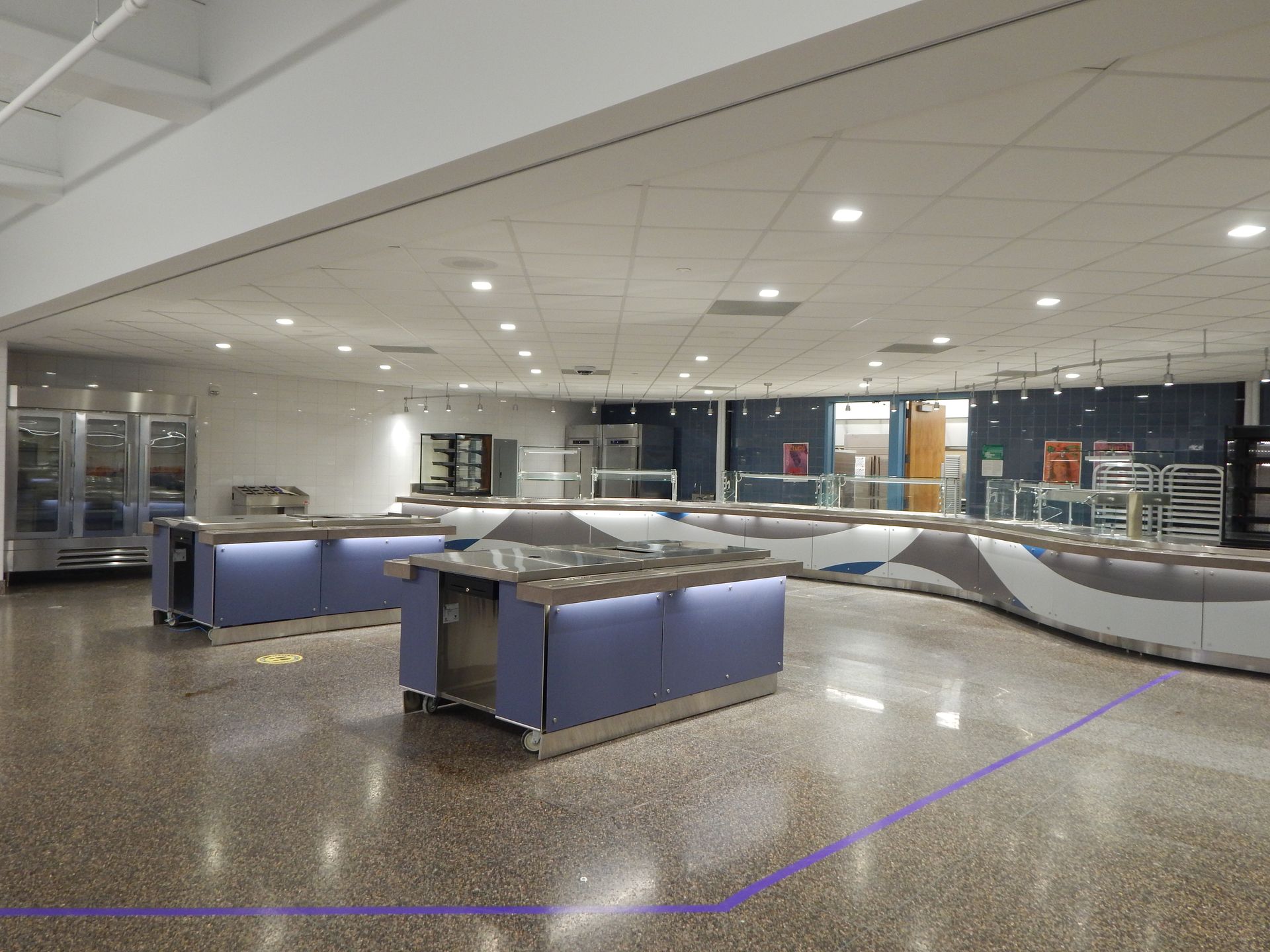Modern cafeteria serving area with blue island counters, stainless steel equipment, and polished speckled floors.