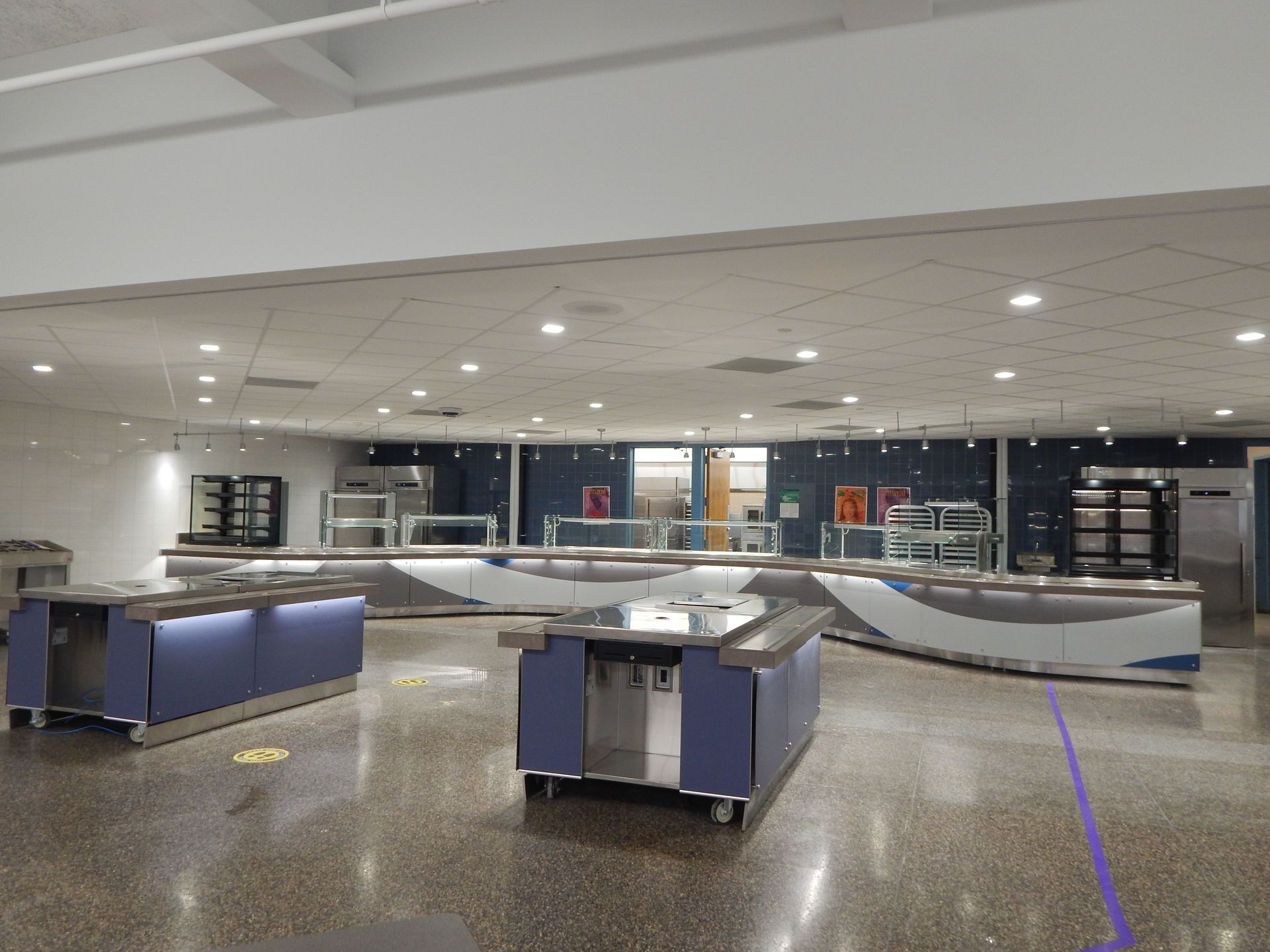 An empty cafeteria serving area with long, blue-paneled food counters and stainless steel surfaces on a polished floor.