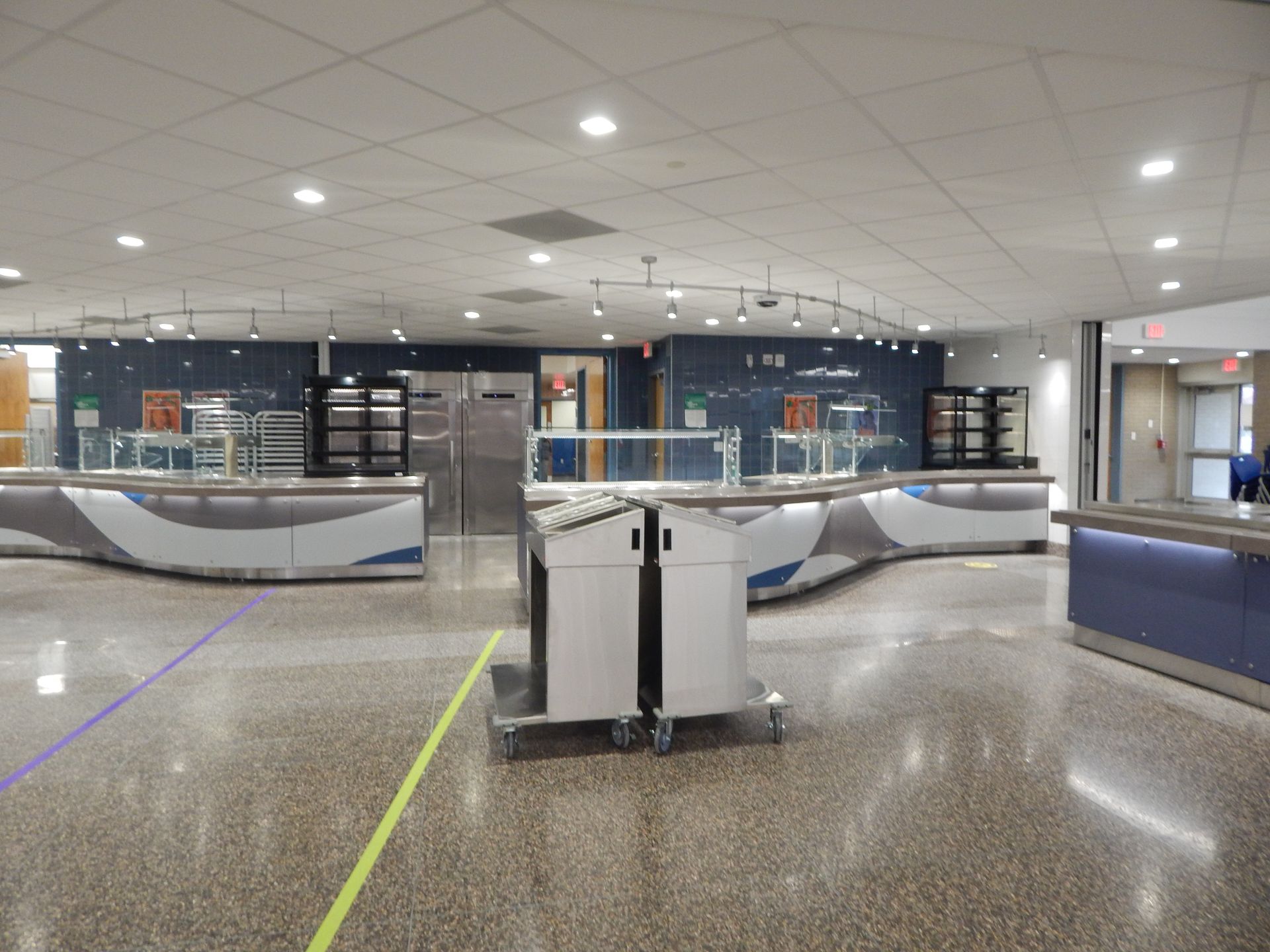 A wide, brightly lit dining hall interior with curved serving counters, a central cart, and blue-accented walls.