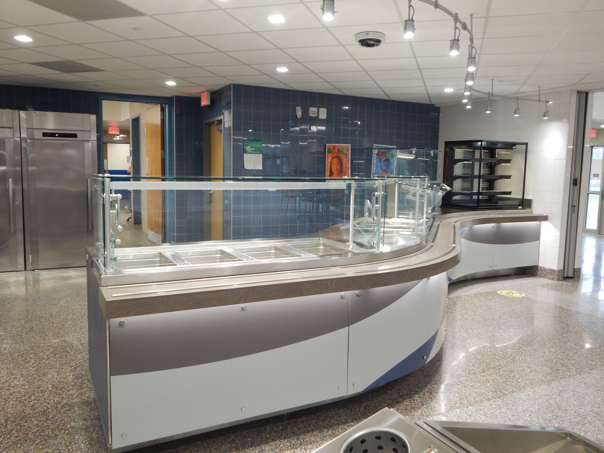 A curved, modern cafeteria serving station with stainless steel food bins, glass sneeze guards, and a gray-white counter.