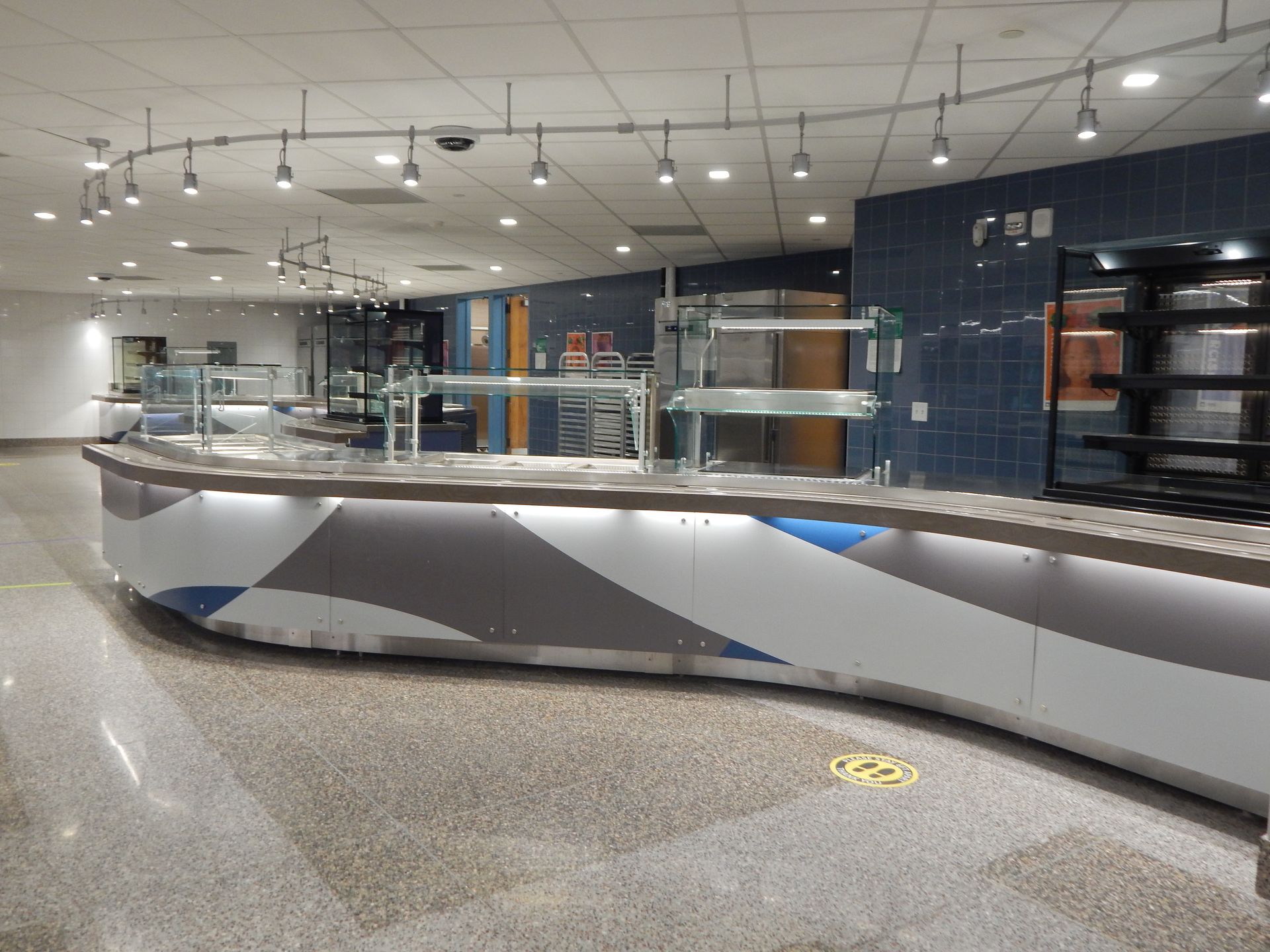 A curving cafeteria serving counter with a white, gray, and blue geometric front, under track lighting on a tiled floor.